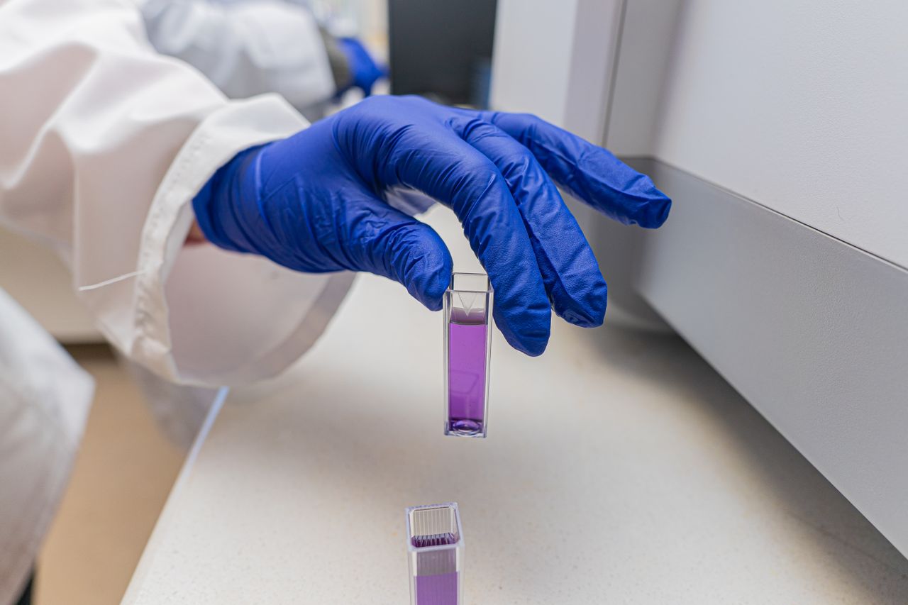 process-img Close-up of a scientist's gloved hand handling a lab sample vial containing a purple liquid.